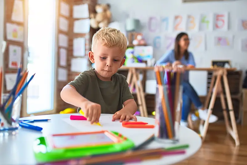 Young Caucasian child playing at playschool with teacher. Child at daycare drawing with color pencils.